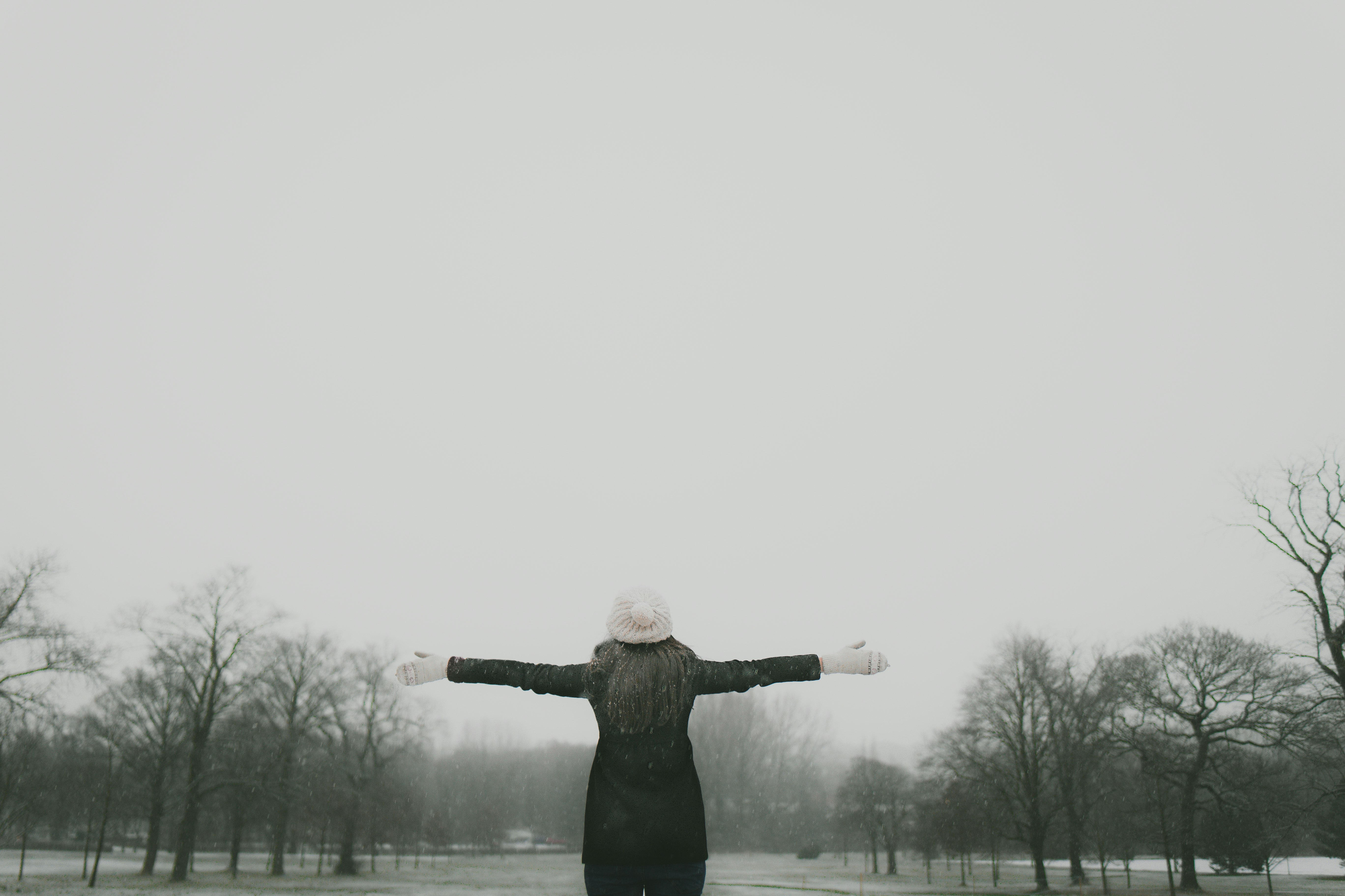 woman in black long-sleeved shirt standing and facing trees with stretched arms under white sky